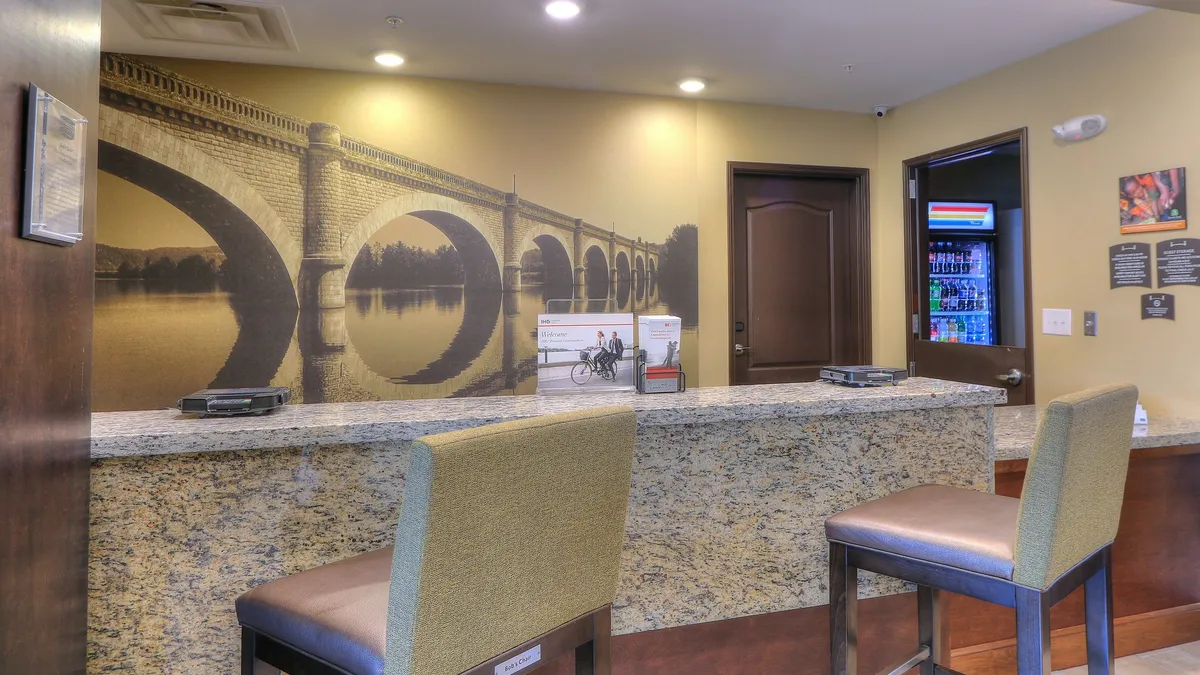 This image captures the hotel's interior reception area, featuring a striking granite desk, a mural that reflects local history, comfortable stool seating, and a vending machine for guest convenience. The scene conveys both professionalism and a welcoming atmosphere.