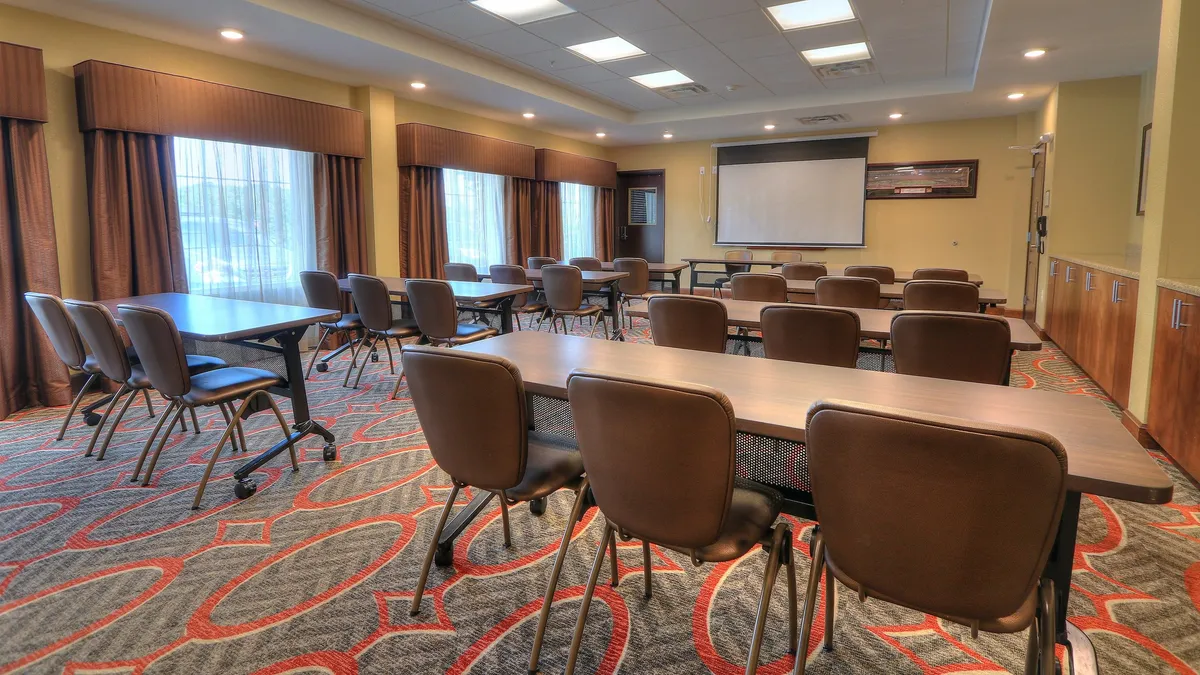 This image showcases a professional, well-lit meeting space with tables arranged for collaboration, large windows filling the room with natural light, and a prominent projector screen at the front. The atmosphere is one of focused productivity, suitable for business meetings, training, or workshops.