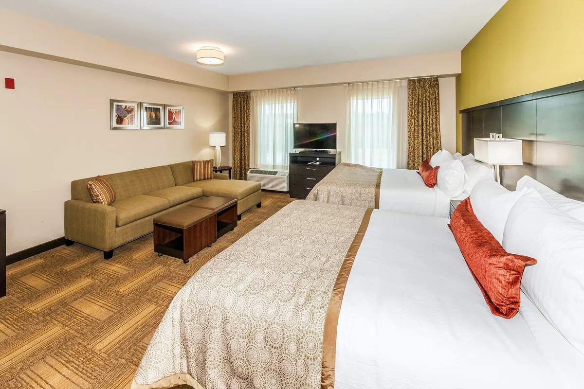 This image captures a bright and inviting studio suite featuring two plush Queen beds and a comfortable sofa seating area. The television and decorative artwork add warmth and unique character, while the layout suggests a space ideal for both relaxation and socializing. The overall mood is cozy, welcoming, and adaptable to various guest needs.