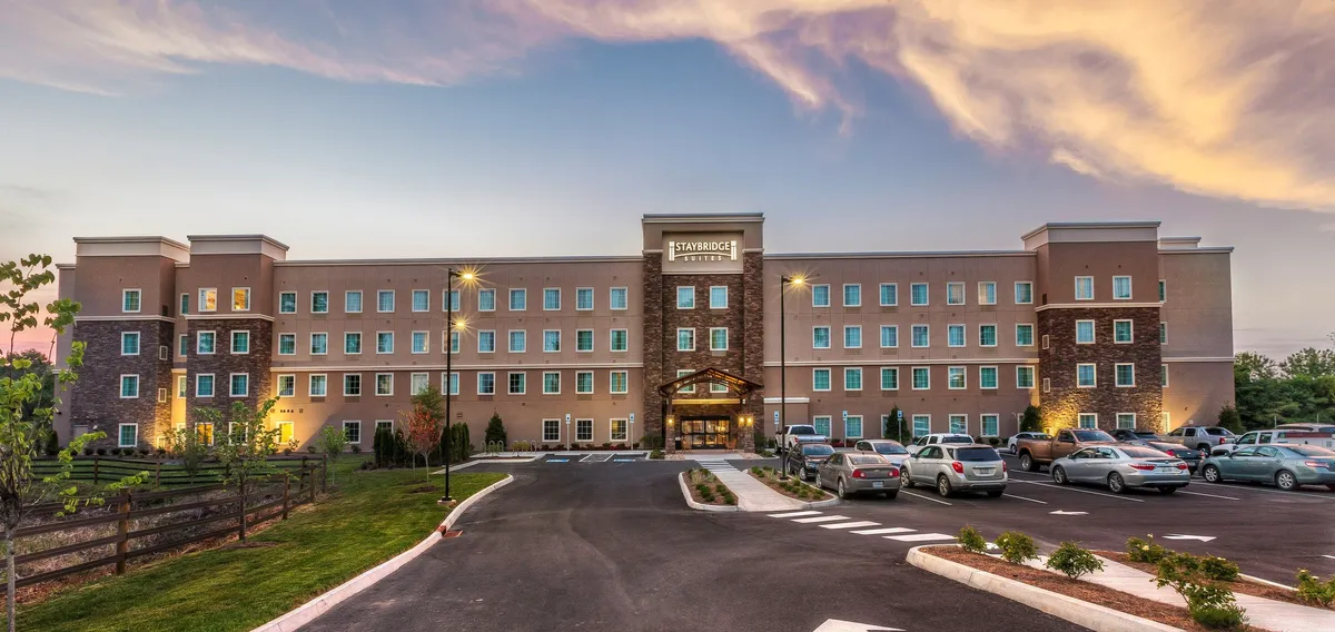 Another exterior perspective of the hotel entrance, highlighting lush landscaping and a spacious parking area. The building glows with soft evening light, evoking feelings of comfort and relaxation at the end of a busy day exploring nearby attractions.