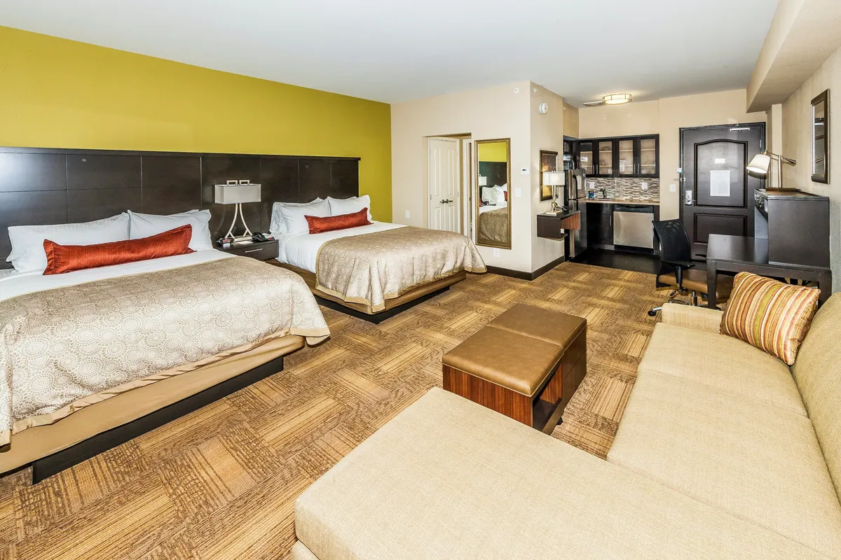 The image showcases a spacious and inviting studio suite featuring two plush queen beds, a cozy sofa seating area perfect for unwinding, a modern desk setup for work or planning, and a sleek TV unit. The room is bathed in warm light, creating a welcoming and comfortable atmosphere ideal for both relaxation and productivity.